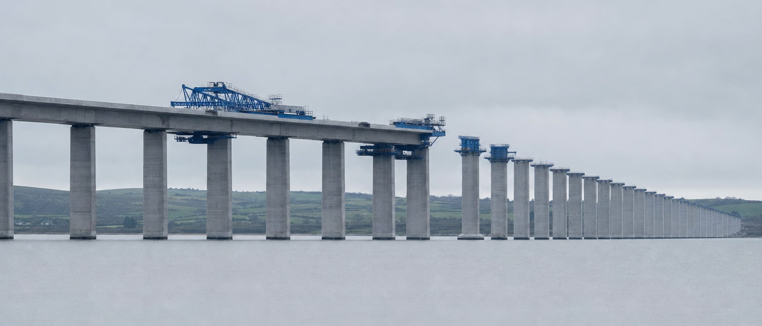 New railway viaduct under construction across an estuary, concrete piers receding into the distance under flat grey light